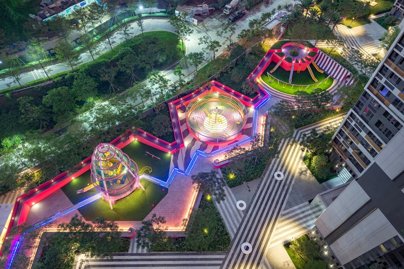 Vue aérienne au crépuscule des aires de jeux multiples des Résidences Buji, reliées par un mur de terrain sinueux peint en rose et violet et parcourues par une « rivière » centrale à LED bleue reliant les espaces de jeux aquatiques.