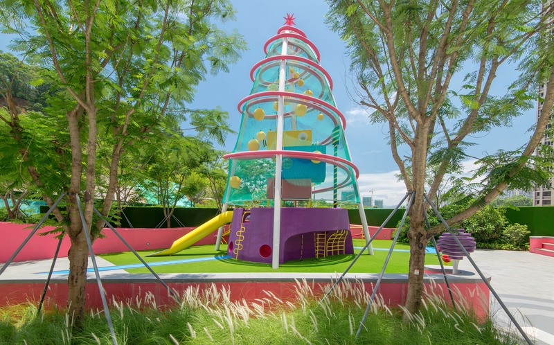 Aire de jeux Playscraper aux Résidences Buji, avec une base violette et des anneaux roses entourant une structure en treillis bleu ponctuée de modules d’escalade jaunes et d’un toboggan, installée au cœur d’une végétation luxuriante sous les tours.