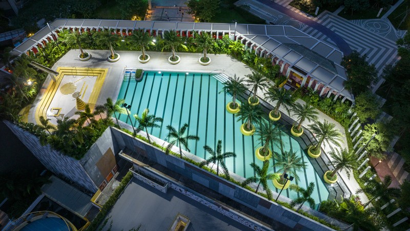 Vue nocturne aérienne de la terrasse-piscine des Résidences Buji, où la piscine de nage turquoise rayée est bordée de nids d’assise circulaires jaunes et de palmiers Washingtonia, et jouxte une piscine pour enfants agrémentée d’éléments ludiques en mosaïq